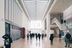 Modern art gallery interior with high glass ceiling, wide hall, people walking, and exhibition signs.