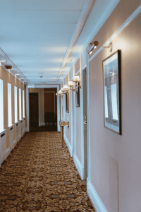 Hotel hallway with patterned carpet, wall lamps, framed pictures, and an exit sign at the end.