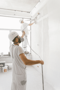 Two construction workers wearing white uniforms, helmets, and respirators spray paint a wall in a bright indoor space.